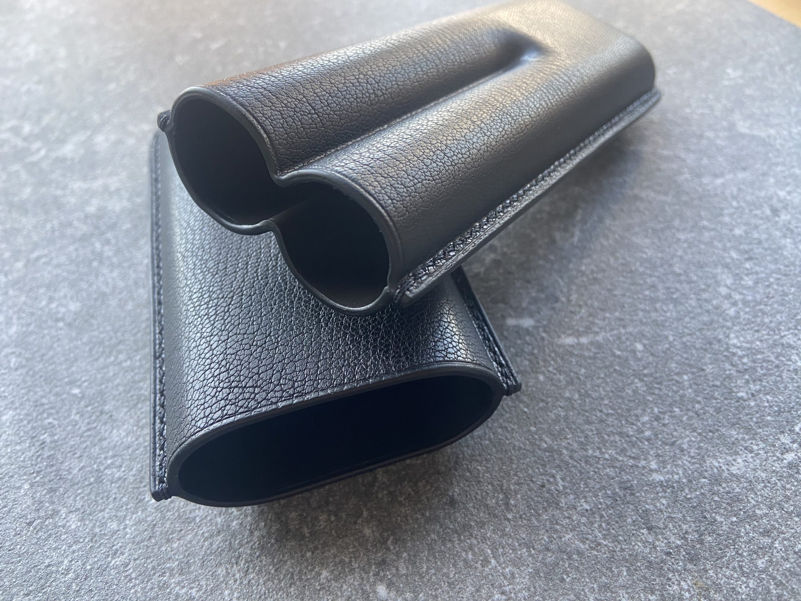 Open black leather cigar case with two rounded compartments on a stone surface
