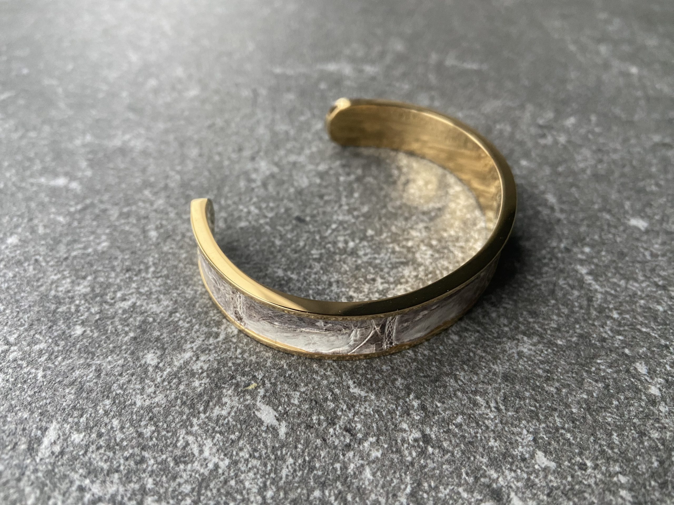 Narrow gold-plated stainless steel cuff bracelet with Himalaya leather inlay on a grey stone background