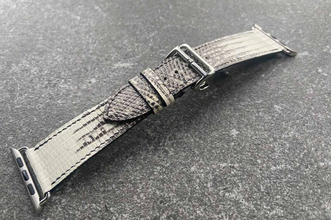 A gray Tegus lizard leather watch band with silver hardware on a textured gray surface