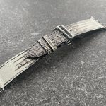 A gray Tegus lizard leather watch band with silver hardware on a textured gray surface