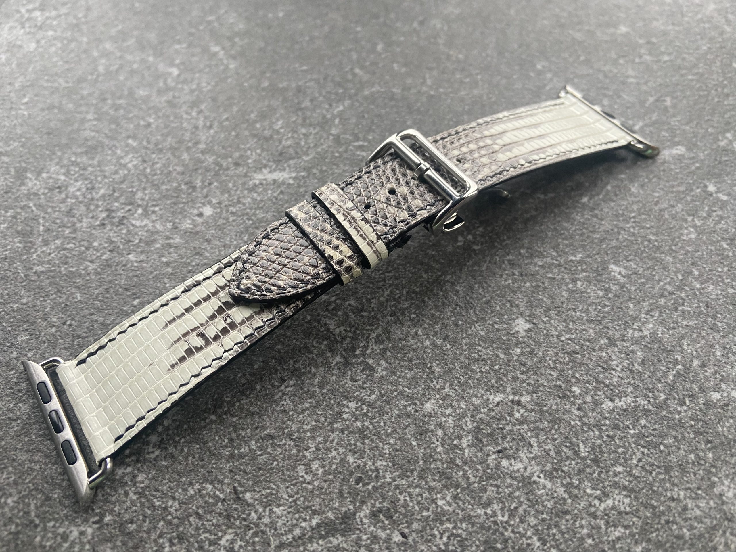 A gray Tegus lizard leather watch band with silver hardware on a textured gray surface