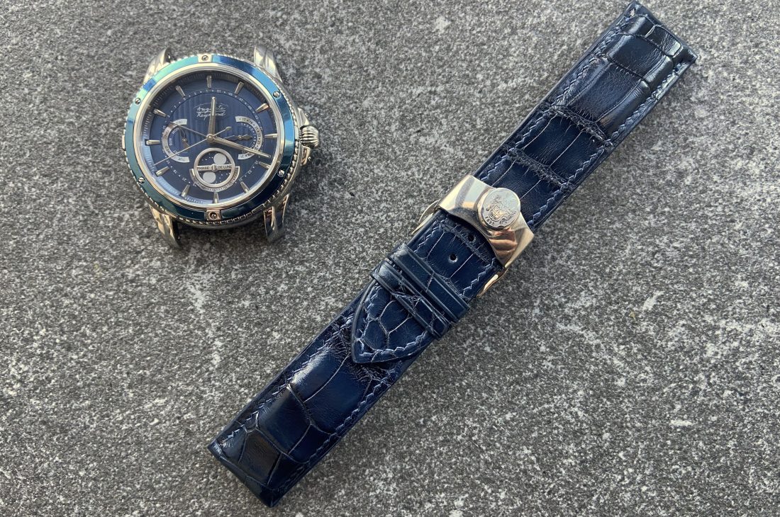 A blue-faced Auguste Reymond Magellan watch is displayed next to a detachable blue leather watch strap on a textured gray background.