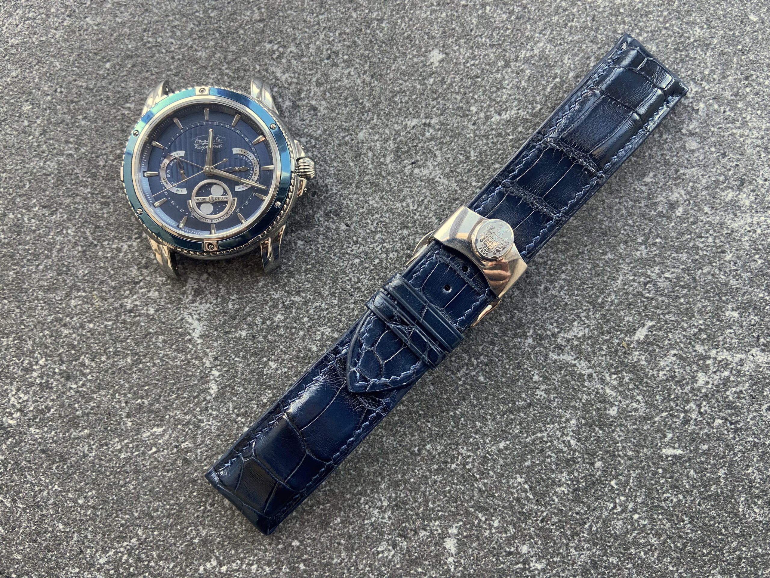 A blue-faced Auguste Reymond Magellan watch is displayed next to a detachable blue leather watch strap on a textured gray background.