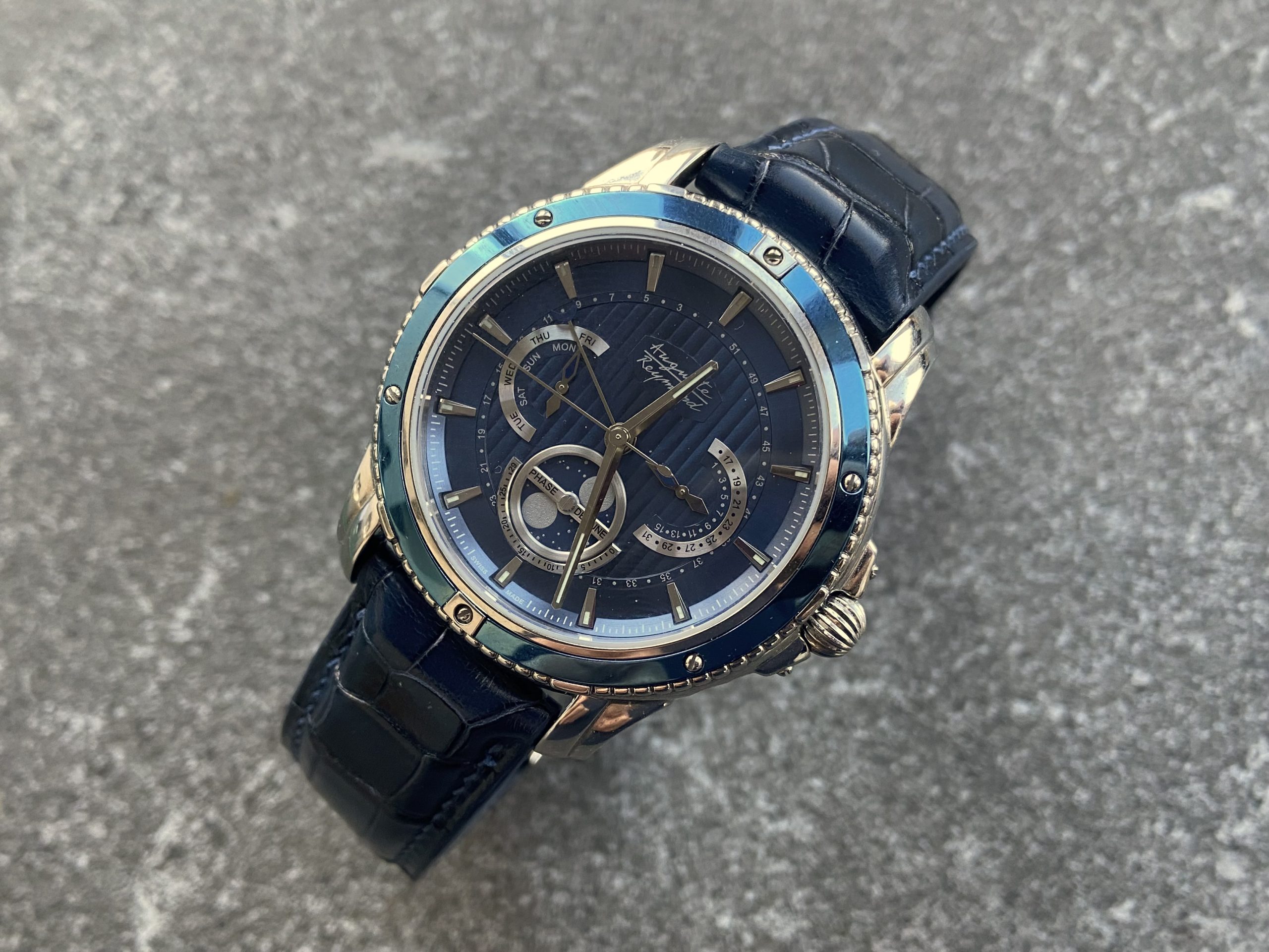 A blue Auguste Reymond Magellan watch with a blue leather strap is displayed on a textured gray background. The watch features a blue dial, a metallic case, and a rotating bezel.