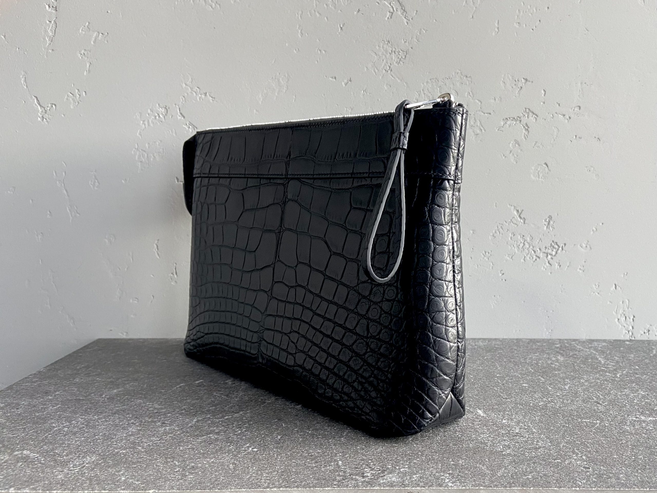Side view of the Pochette Voyage Souple in black alligator leather with wrist strap