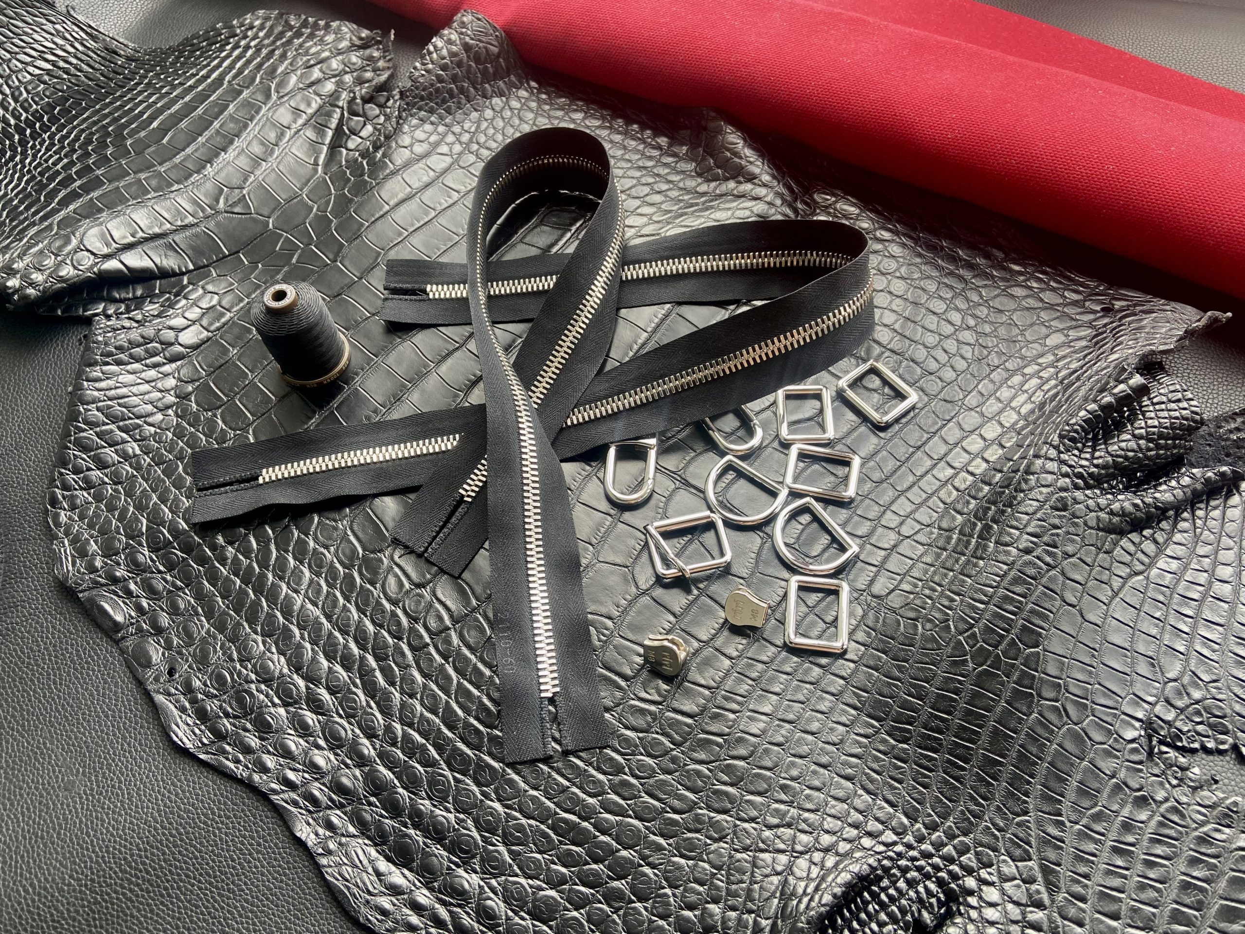 Selected materials for bespoke travel bag including matte black alligator leather, RiRi zippers, thread, hardware, and red Oscar lining