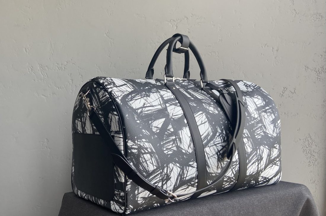 Bespoke duffle bag in brushstroke-patterned leather with black handles and detachable shoulder strap, placed on dark cloth background.