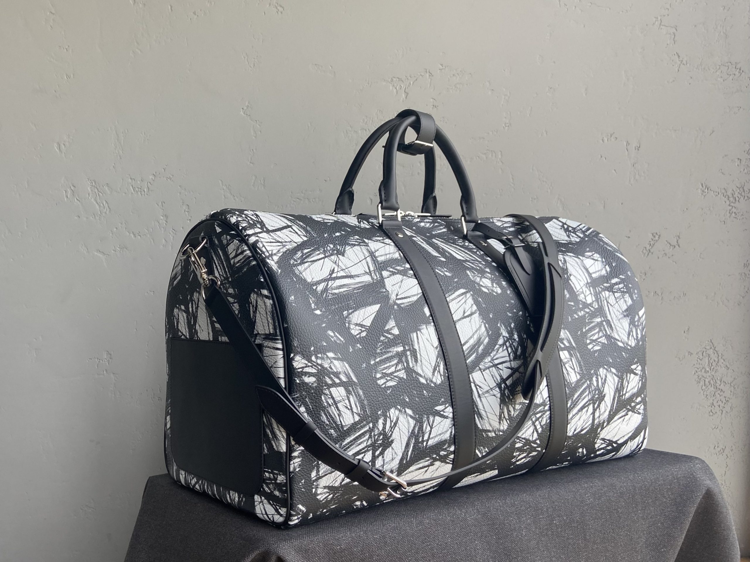 Bespoke duffle bag in brushstroke-patterned leather with black handles and detachable shoulder strap, placed on dark cloth background.