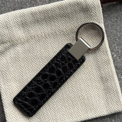 Black structured grain leather key fob with polished 316L stainless steel hardware on a light herringbone fabric pouch