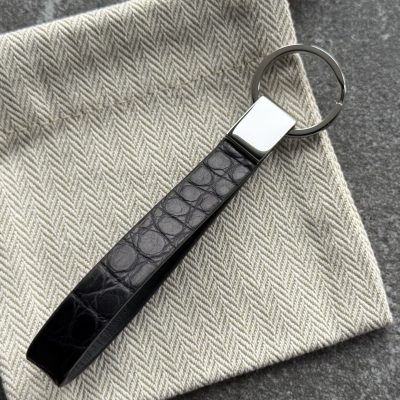 Black structured grain leather loop key holder with polished 316L stainless steel hardware on a light herringbone fabric pouch