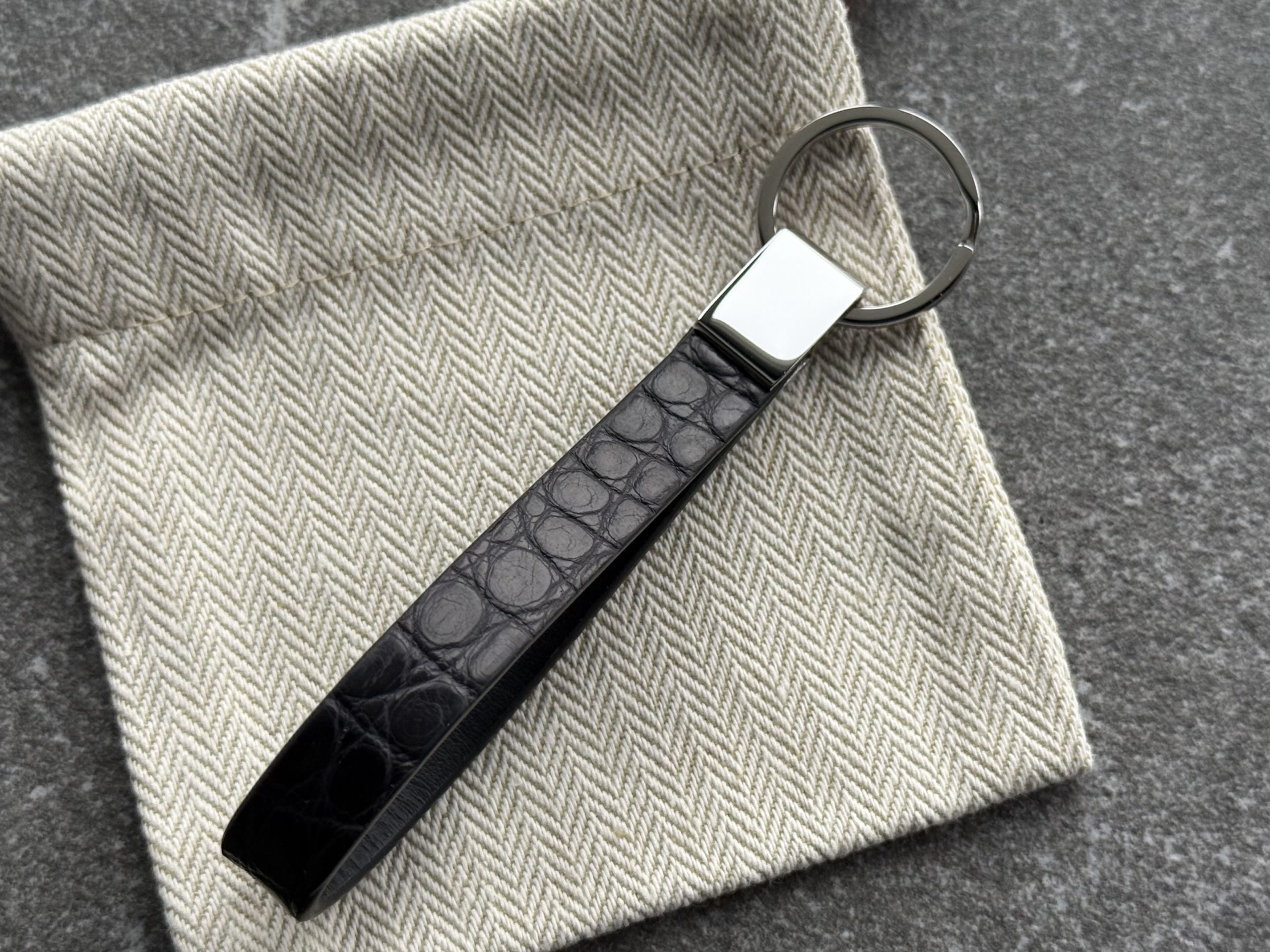 Black structured grain leather loop key holder with polished 316L stainless steel hardware on a light herringbone fabric pouch