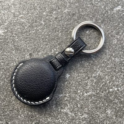 Leather keychain for Apple AirTag in black leather with white contrast stitching shown from the back on a stone surface