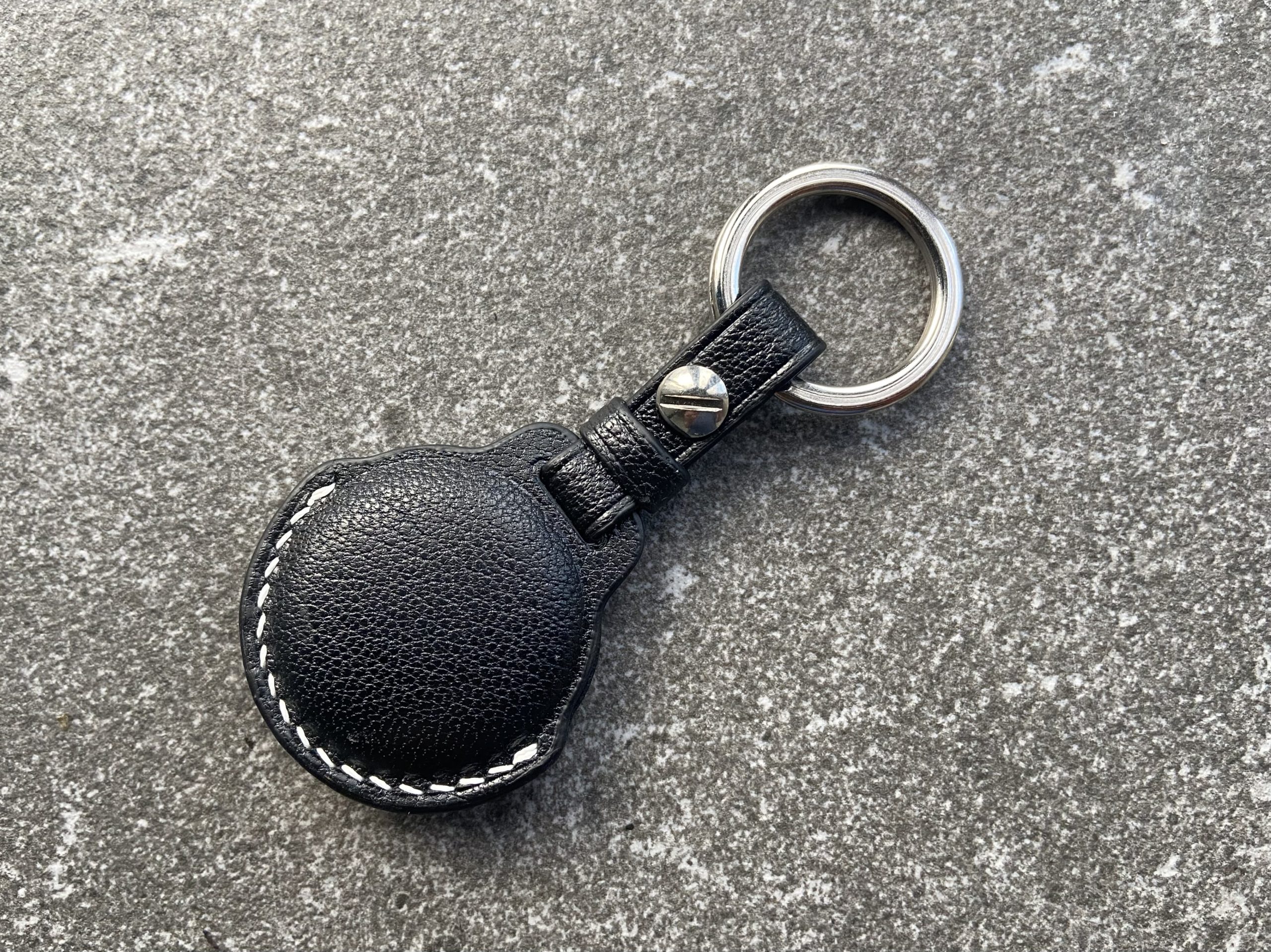 Leather keychain for Apple AirTag in black leather with white contrast stitching shown from the back on a stone surface